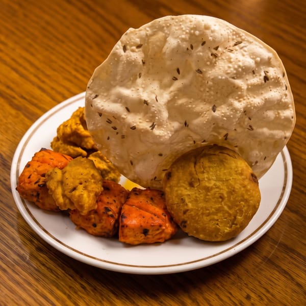Plate of Indian appetizers with papadum, orange tikka, yellow pakoras, and a golden fried patty.