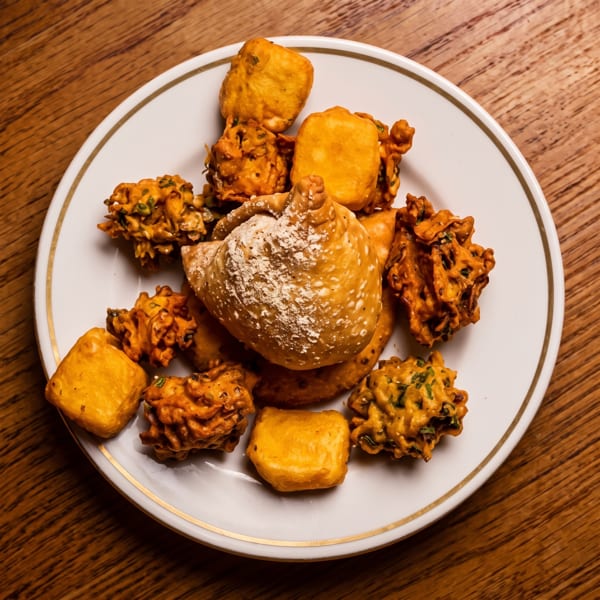 A white plate filled with golden fried samosas, paneer pakoras, and vegetable pakoras on a wooden table.