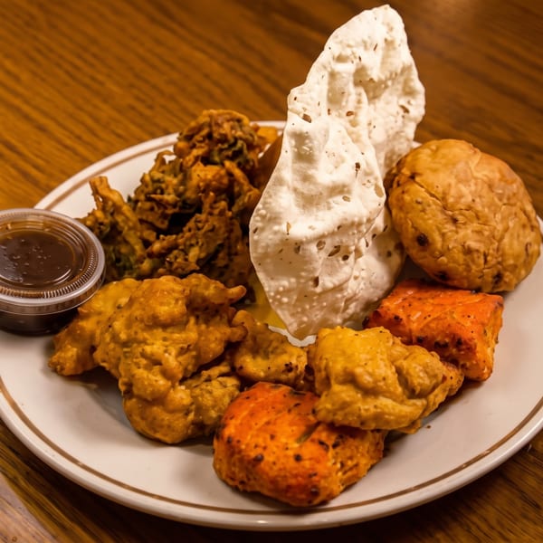 Plate of various Indian appetizers including pakoras, paneer tikka, samosas, papadum, and dipping sauce.