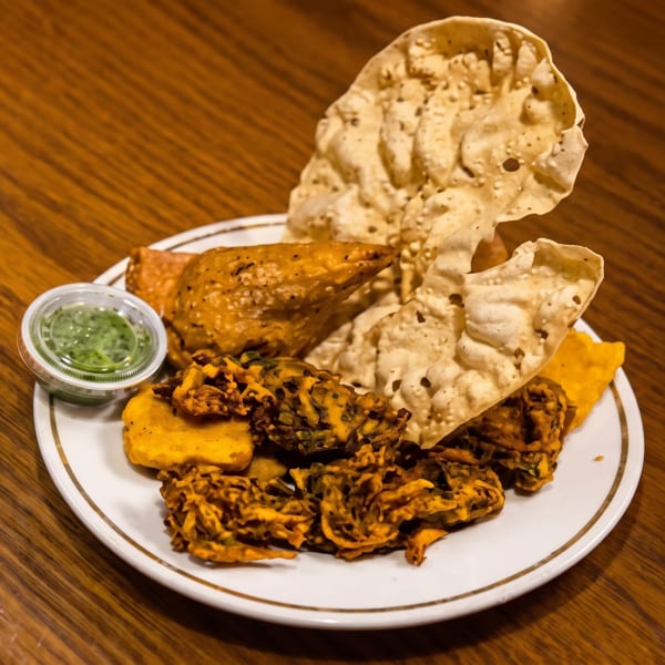 A white plate holds crispy papadum, golden samosas, pakoras, and green chutney on a wooden table.